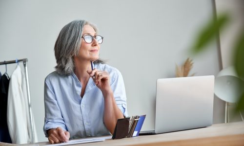 Mature woman with glasses at a desk with a laptop, gazing thoughtfully