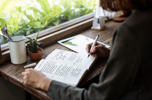 Person writing in a journal at a rustic wooden desk beside a window