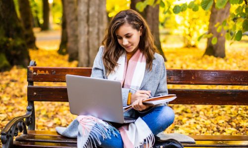 Woman on a park bench in autumn working on a laptop with a notebook