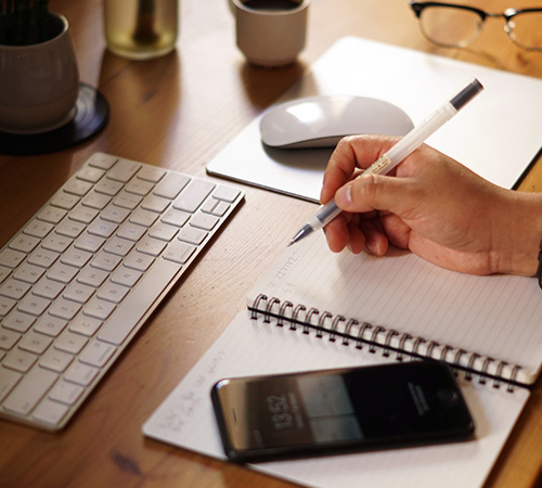 Hand writing in a spiral notebook on a desk with a keyboard and phone